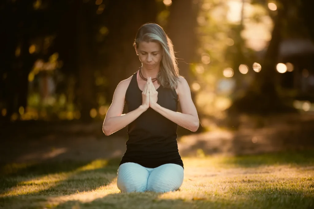 Michelline meditando ao ar livre, com as mãos em prece, na luz dourada do fim de tarde